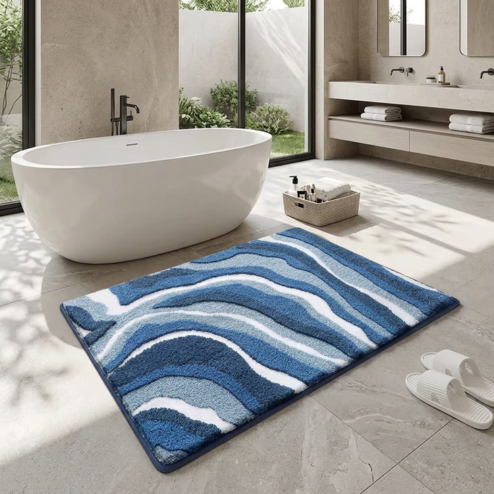 A rectangular bath mat with a wavy abstract pattern in shades of blue, navy, and white, placed on a light gray tiled bathroom floor next to a white freestanding bathtub.