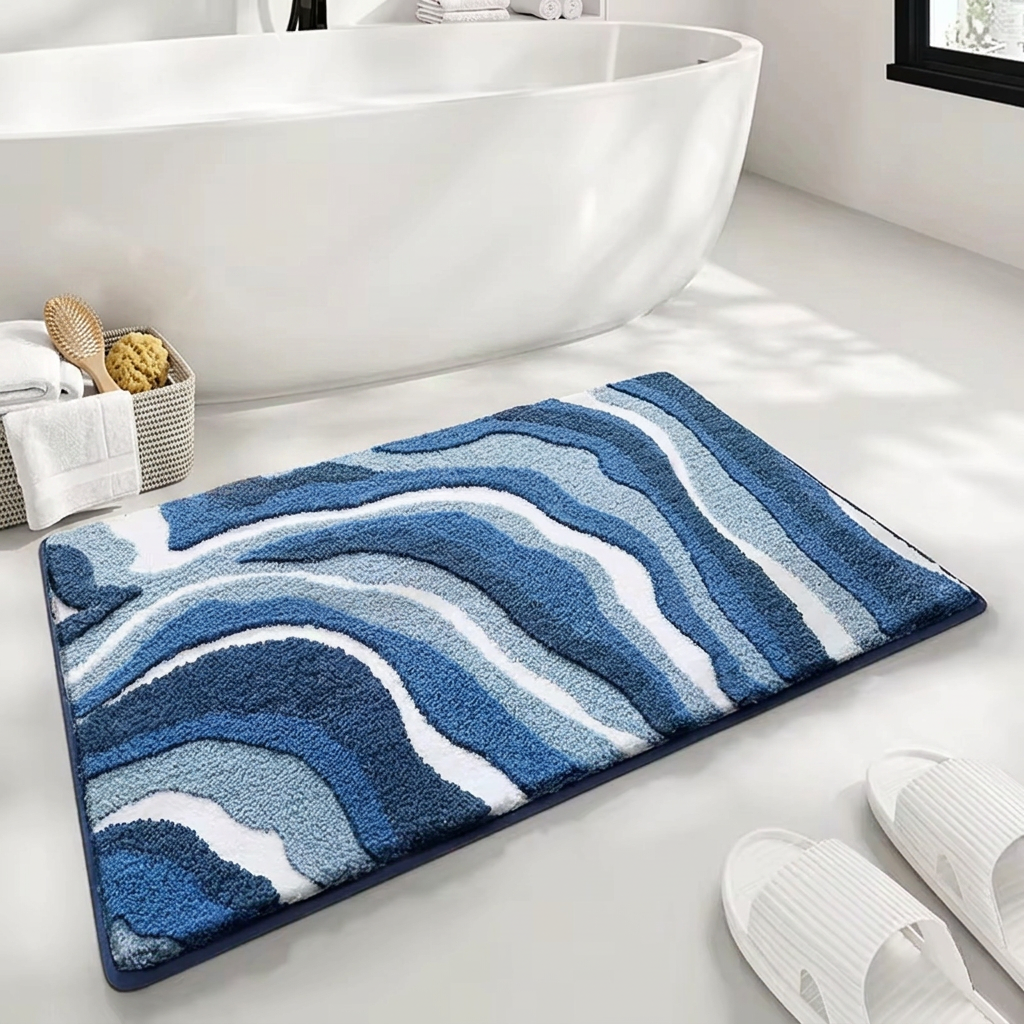 A rectangular bath mat with a wavy, layered abstract pattern in shades of blue, navy, and white, styled on a white bathroom floor next to a bathtub, white slippers, and a small basket of bath accessories.