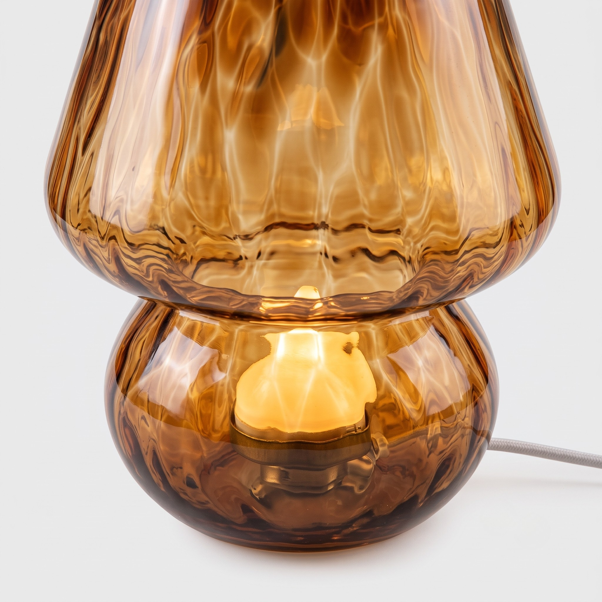 A close-up of a modern amber glass table lamp with a textured, wavy design, lit from within to show a warm yellow glow, on a light gray background.