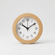 A minimalist round analog clock with a light natural wood frame, a white face, and black numerals and hands, photographed against a seamless light gray background.
