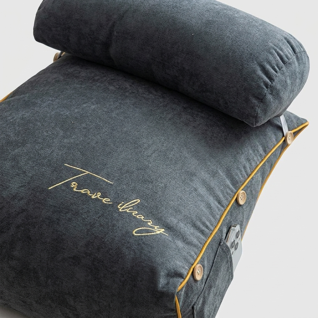 Dark gray travel pillow with a matching rolled headrest, featuring gold piping, decorative buttons, and gold script embroidery reading 'Travel library' on a light gray background.