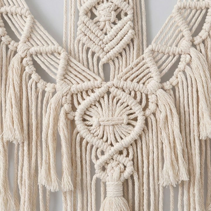 Cream macrame wall hanging with intricate woven geometric patterns and long fringe tassels, shown against a light neutral background.