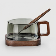 A modern glass mug with a translucent smoky gray body, a square wooden handle, and a matching wooden spoon resting inside, all set on a round wooden base against a light gray background.