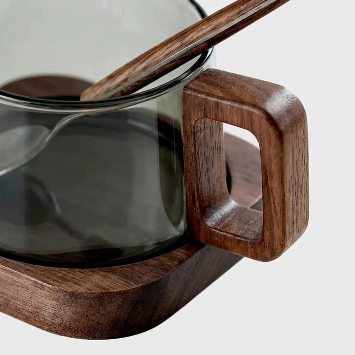 A close-up of a dark smoky glass mug with a thick, square-shaped wooden handle and matching wooden base, featuring a wooden spoon resting inside.