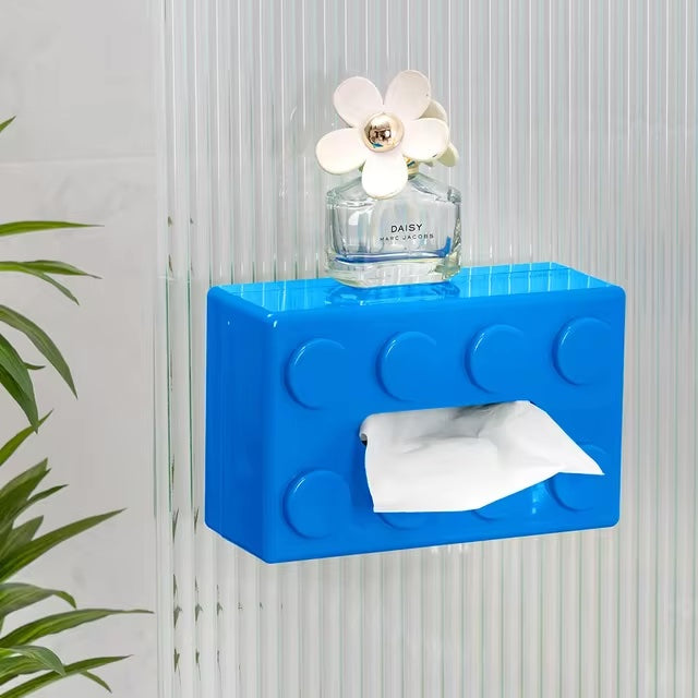 Blue brick-shaped tissue holder with a white flower and perfume bottle on a light gray wall.
