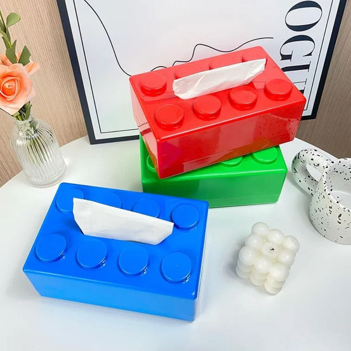 Three colorful tissue boxes (red, blue, green) on a surface with a vase and decorative items in the background.