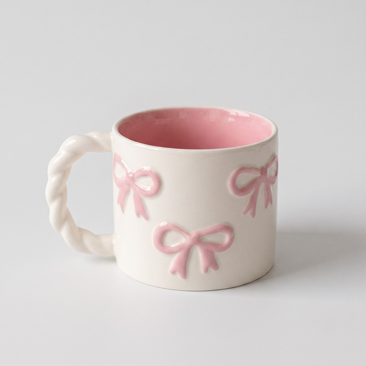 A white ceramic mug with a twisted rope-style handle, featuring embossed pink bows on the exterior and a pink glazed interior, set against a plain light gray background.