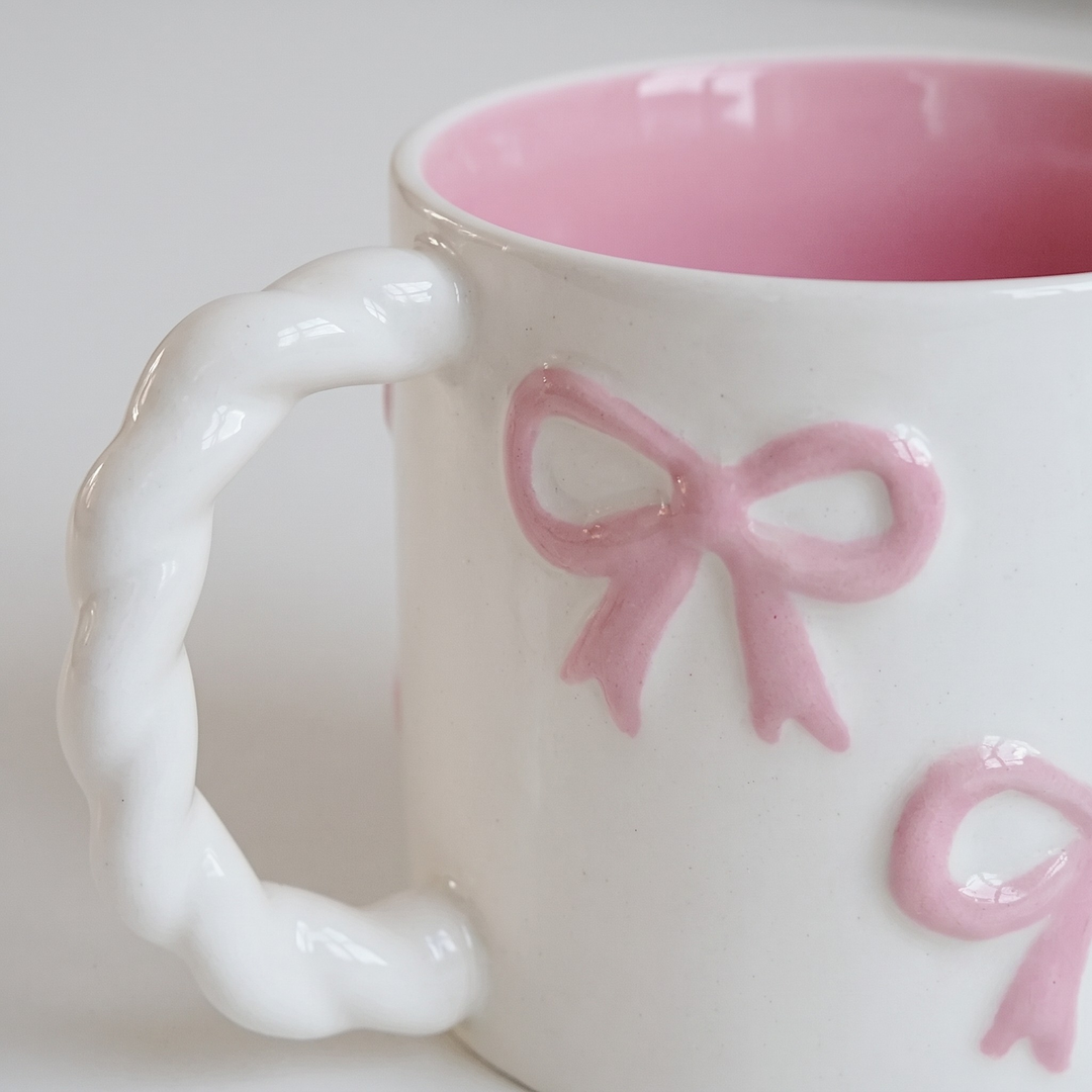 A close-up of a white ceramic mug with a twisted handle, featuring embossed pink ribbon bows on the exterior and a pink interior.