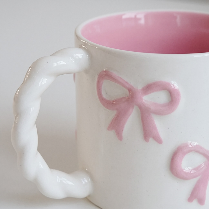 A close-up of a white ceramic mug with a twisted handle, featuring embossed pink ribbon bows on the exterior and a pink interior.