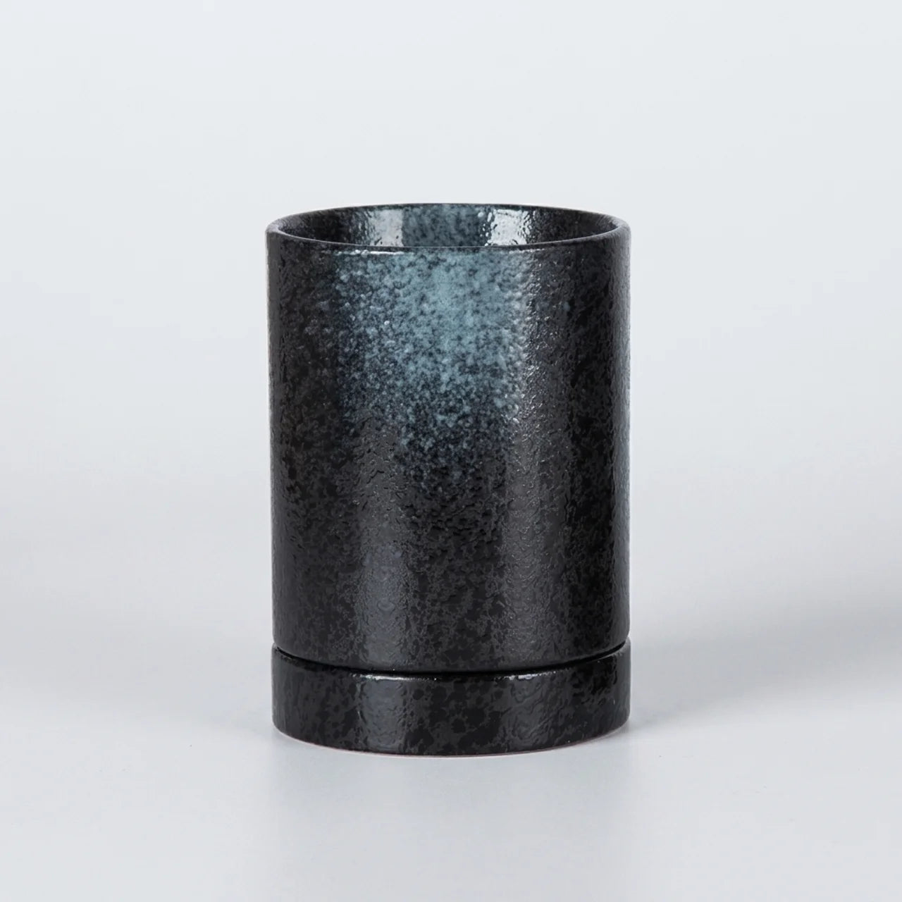 A cylindrical black ceramic cup or candle holder with a textured, metallic-like finish and a flared base, shown on a light gray background.