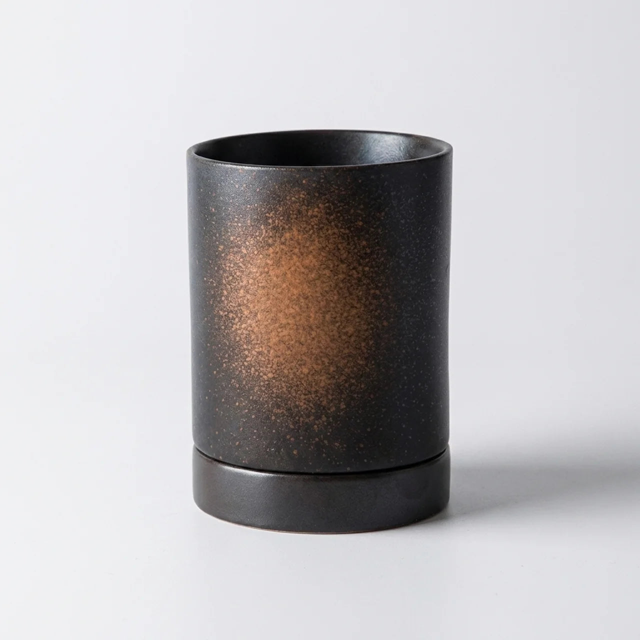 A matte black cylindrical cup or candle holder with a textured metallic bronze-gold speckled pattern on the front, set against a clean light gray background.