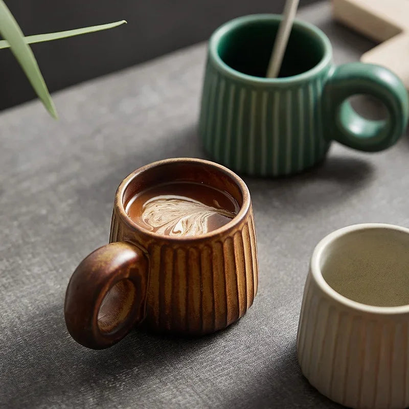 Three ceramic mugs in different colors on a gray surface with a plant in the background.