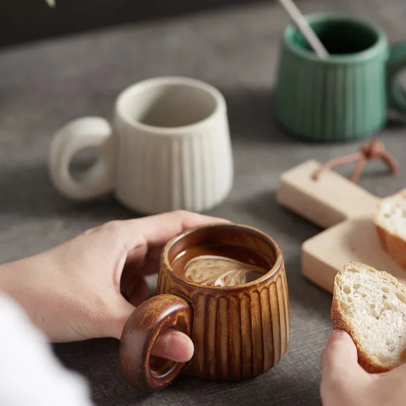 Person holding a brown ceramic mug with a drink, surrounded by other mugs and bread on a table.
