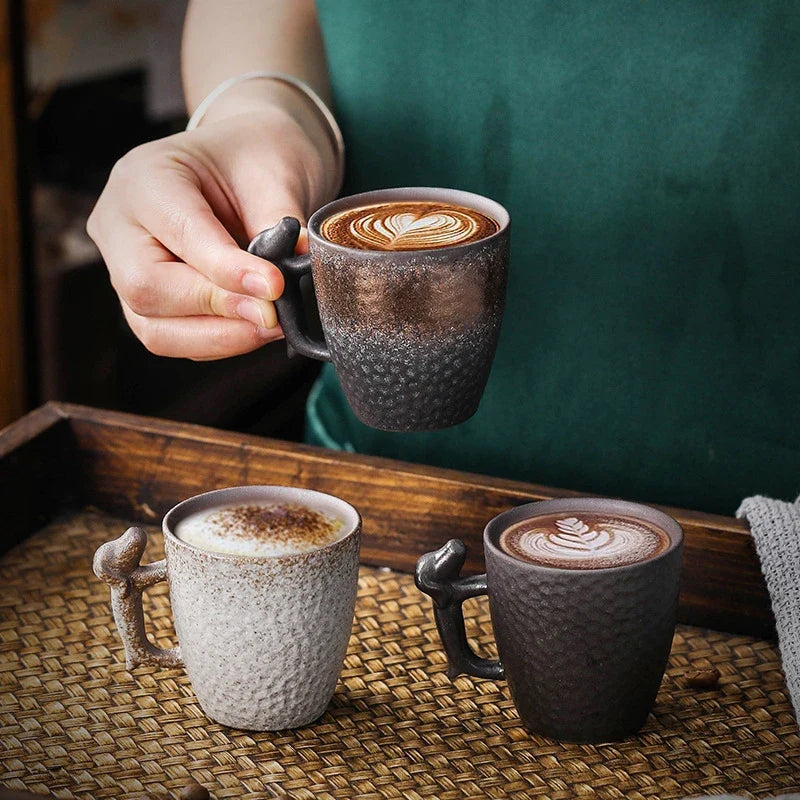 Person holding a textured mug with a cappuccino, surrounded by three other mugs on a woven surface.