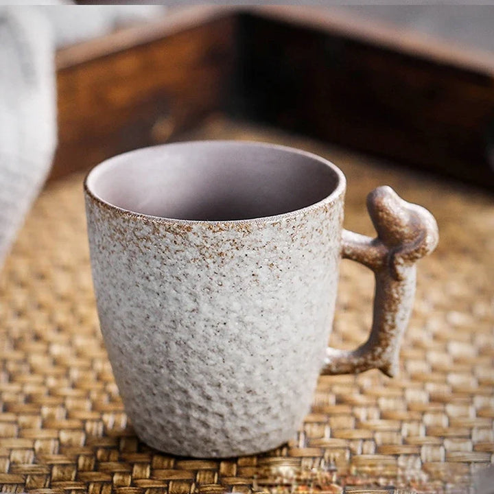 A speckled ceramic mug with a textured, rustic finish and a sculptural handle, resting on a woven straw mat.