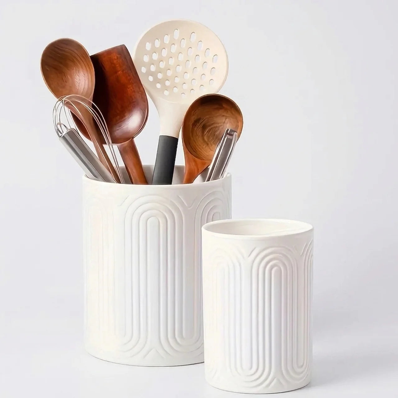 A white ceramic kitchen utensil holder with a matching cup, filled with wooden and metal kitchen tools, on a light gray background.