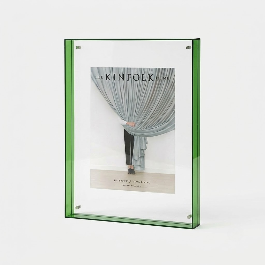 A green-framed vertical picture frame holding a magazine-style insert with a photograph of draped curtains and the text 'THE KINFOLK HOME', shown on a clean white background.