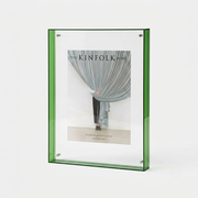 A green-framed vertical picture frame holding a magazine-style insert with a photograph of draped curtains and the text 'THE KINFOLK HOME', shown on a clean white background.