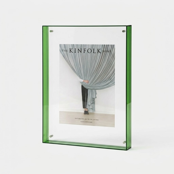 A green-framed vertical picture frame holding a magazine-style insert with a photograph of draped curtains and the text 'THE KINFOLK HOME', shown on a clean white background.
