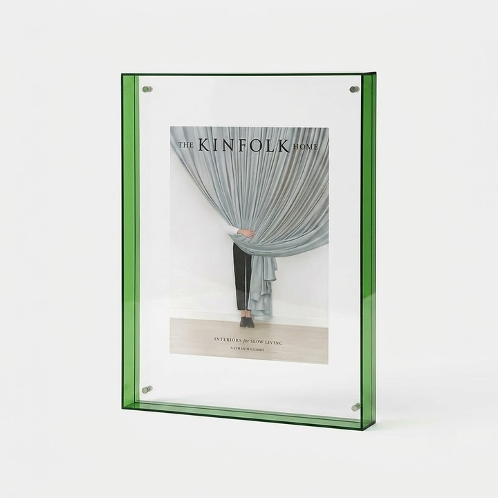 A green-framed vertical picture frame holding a magazine-style insert with a photograph of draped curtains and the text 'THE KINFOLK HOME', shown on a clean white background.