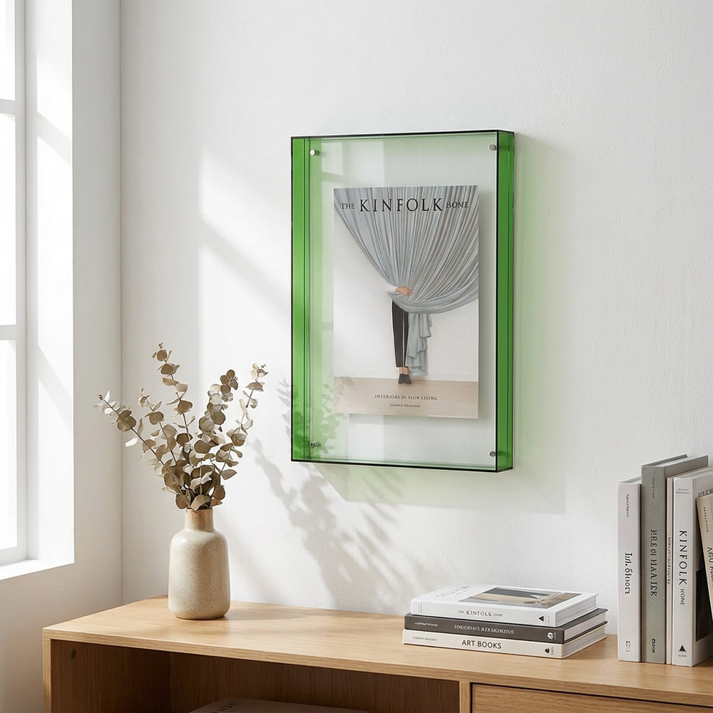 A rectangular acrylic picture frame with a translucent green border hangs on a white wall above a wooden console table. The frame contains a fashion magazine cover, and the scene is styled with a beige vase of dried eucalyptus and books on the table.