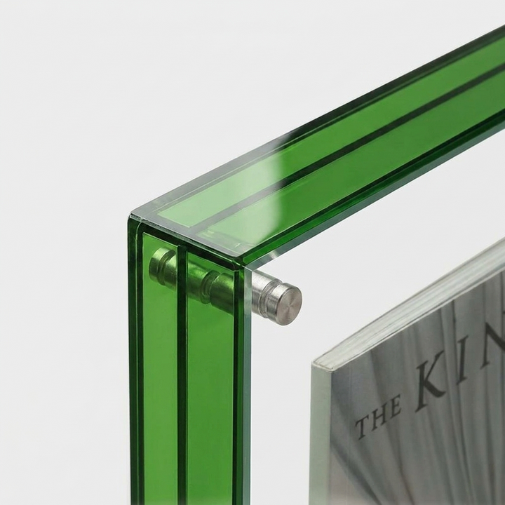 Close-up of a green transparent acrylic or glass frame corner with a silver metal fastener, holding a magazine against a light gray background.