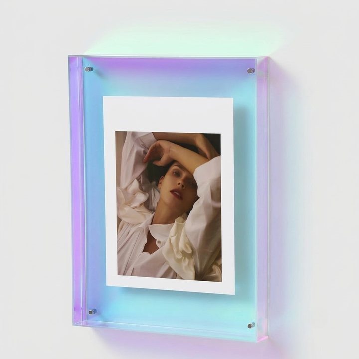 A modern acrylic picture frame with a built-in LED light bar, casting a soft purple and blue glow. The frame is mounted on a plain white wall and contains a sample portrait photo with a white border.