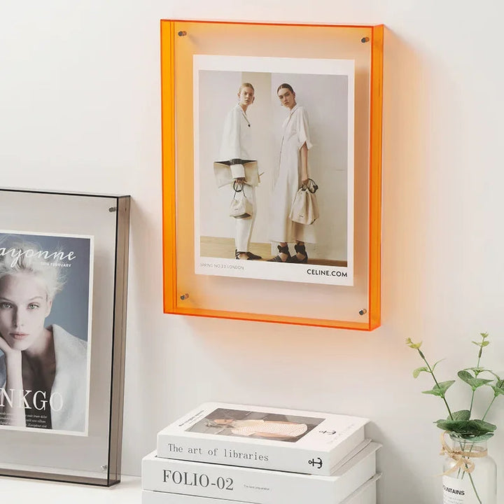 Framed picture of two women in white outfits with a Celine logo, placed on a shelf with books and a plant.