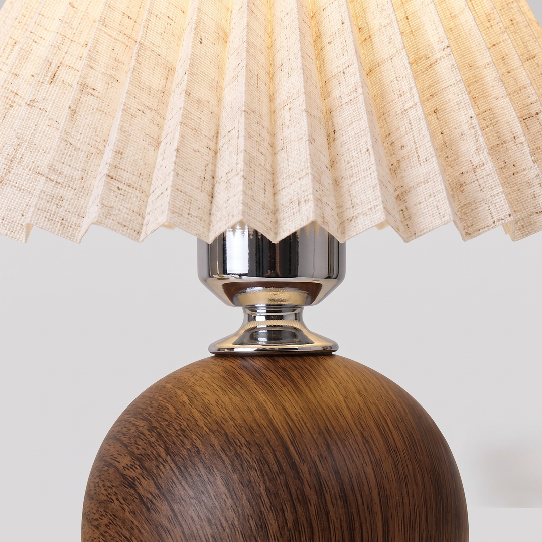 A close-up of a table lamp featuring a pleated beige woven shade, a polished silver metal neck, and a round dark brown wooden base.