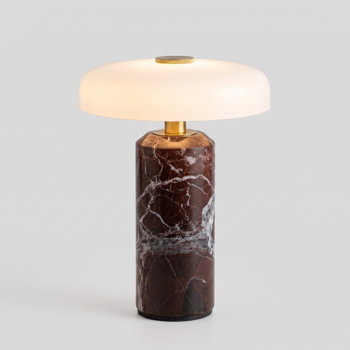 A modern table lamp with a cylindrical dark brown and black marble base featuring white veining, topped with a gold-finished collar and a glowing, dome-shaped frosted white shade.