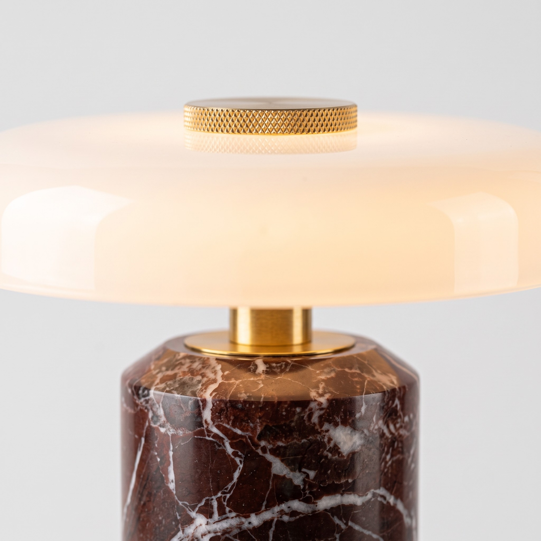 A close-up of a table lamp with a warm white oval glass shade, a textured gold metal top, and a dark brown marble base with white veining.