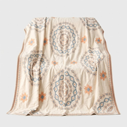 A cream-colored throw blanket with a bohemian-style pattern featuring blue and orange medallions and starburst motifs, draped against a light gray background.