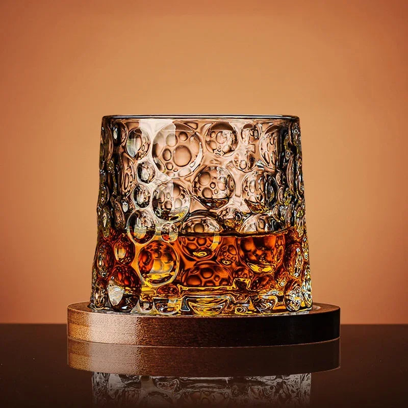 Glass with bubble design filled with a amber liquid on a wooden coaster against an orange background