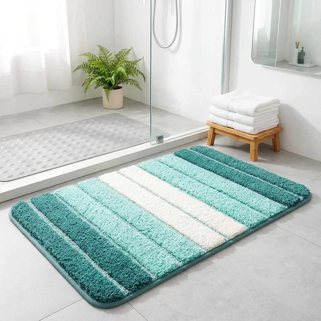 A rectangular bath mat with vertical striped patterns in shades of teal, mint green, and white, placed on a light gray tiled bathroom floor next to a glass shower door.
