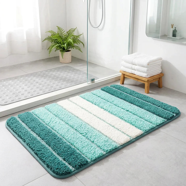 A rectangular bath mat with vertical striped patterns in shades of teal, mint green, and white, placed on a light gray tiled bathroom floor next to a glass shower door.