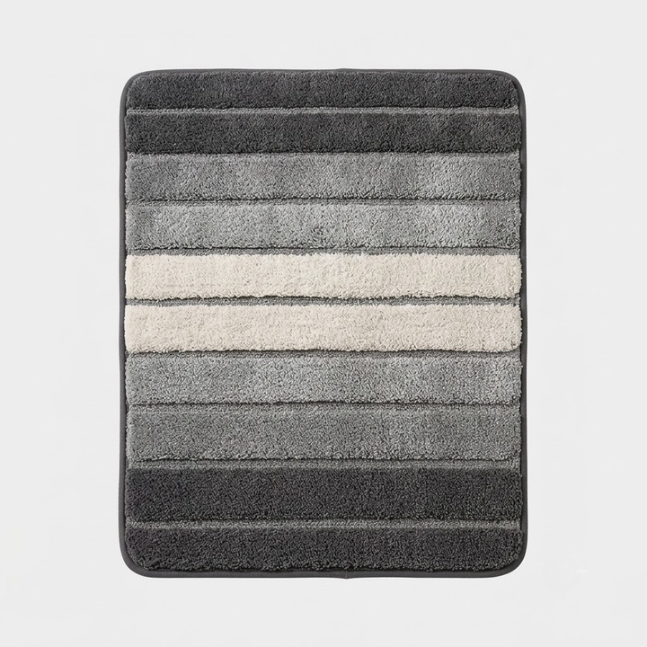 A rectangular bath mat with rounded corners, featuring horizontal striped texture in shades of dark gray, medium gray, and a central off-white stripe, shown against a light gray background.