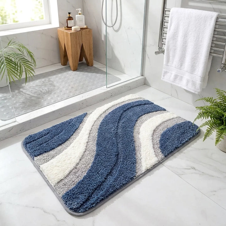 A rectangular bath mat with a wavy, layered shaggy pattern in shades of blue, gray, and white, placed on a light marble bathroom floor next to a glass shower door.