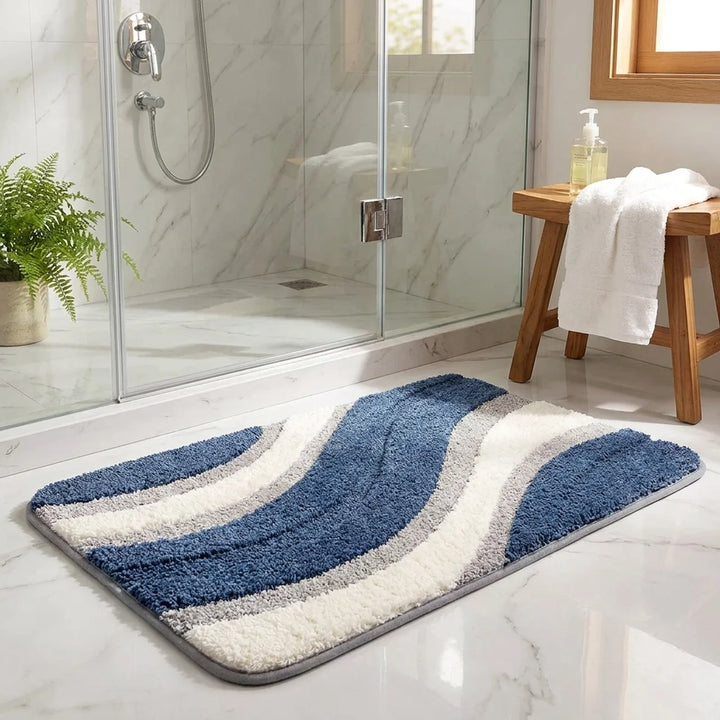 A rectangular bath mat with a wavy striped pattern in navy blue, white, and light gray, placed on a white marble bathroom floor in front of a glass shower door.