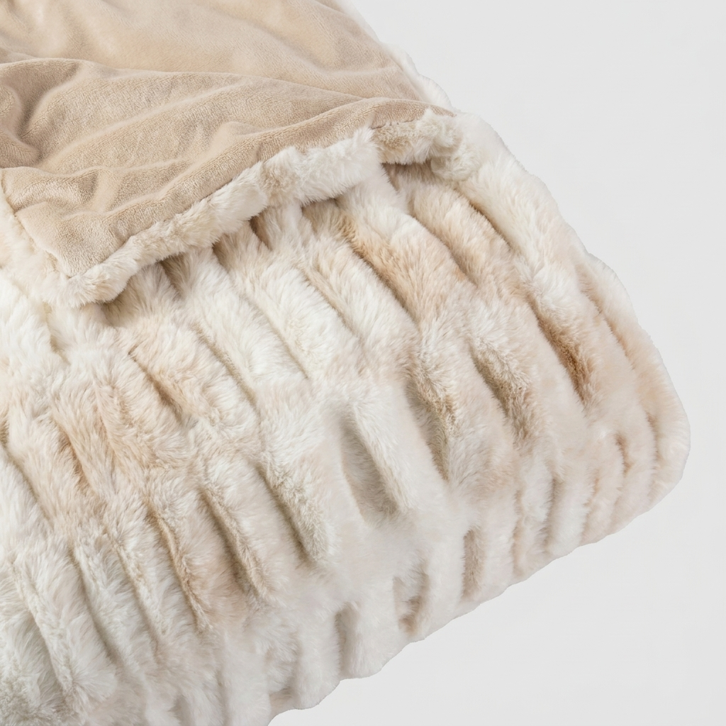 A close-up of a folded plush throw blanket showing two textures: a ribbed faux-fur style in cream and a smooth suede-like section in light beige, set against a plain light gray background.