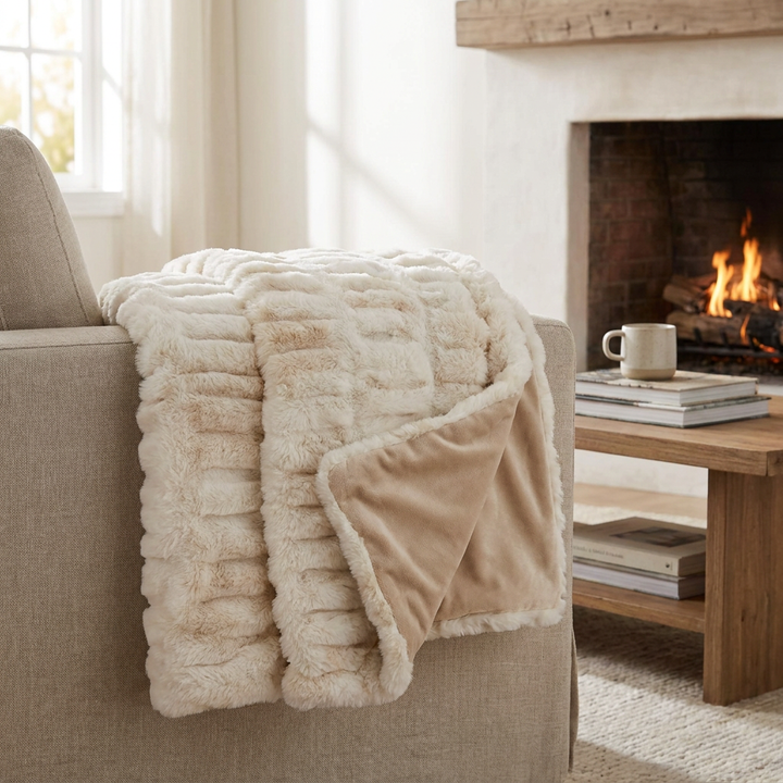 A cozy cream and beige textured throw blanket draped over a beige armchair in a living room setting with a burning fireplace and a wooden coffee table nearby.