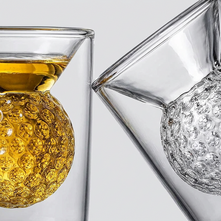 Two clear glass tumblers with sculptural textured globes beneath the rims, one filled with amber liquid and the other empty, shown on a light gray background.