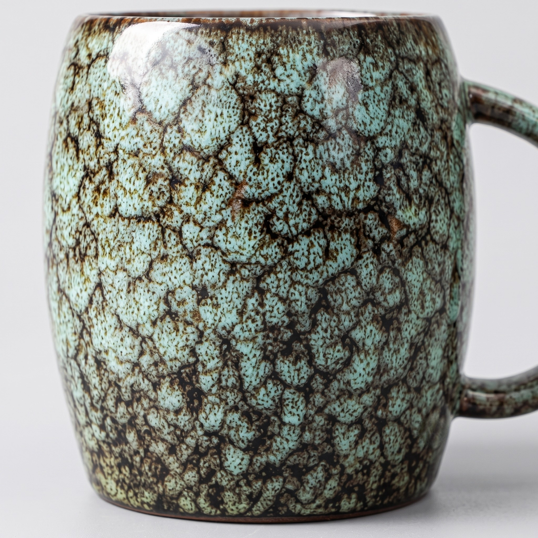 A ceramic mug with a textured, crackled glaze in light turquoise and dark brown, shown against a neutral light gray background.