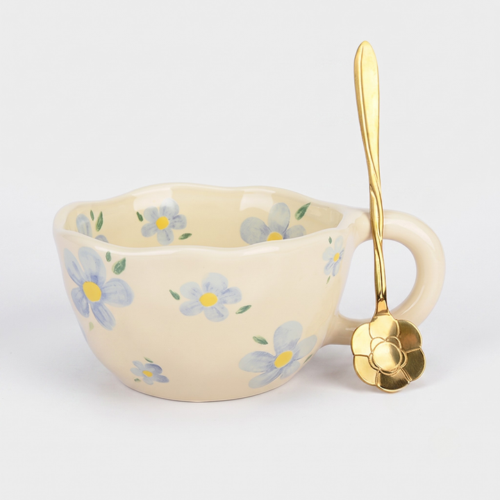 A cream-colored ceramic cup with a handle, decorated with a pattern of light blue and yellow forget-me-not flowers with green leaves. A matching gold spoon with a flower-shaped bowl rests against the cup on a light gray background.
