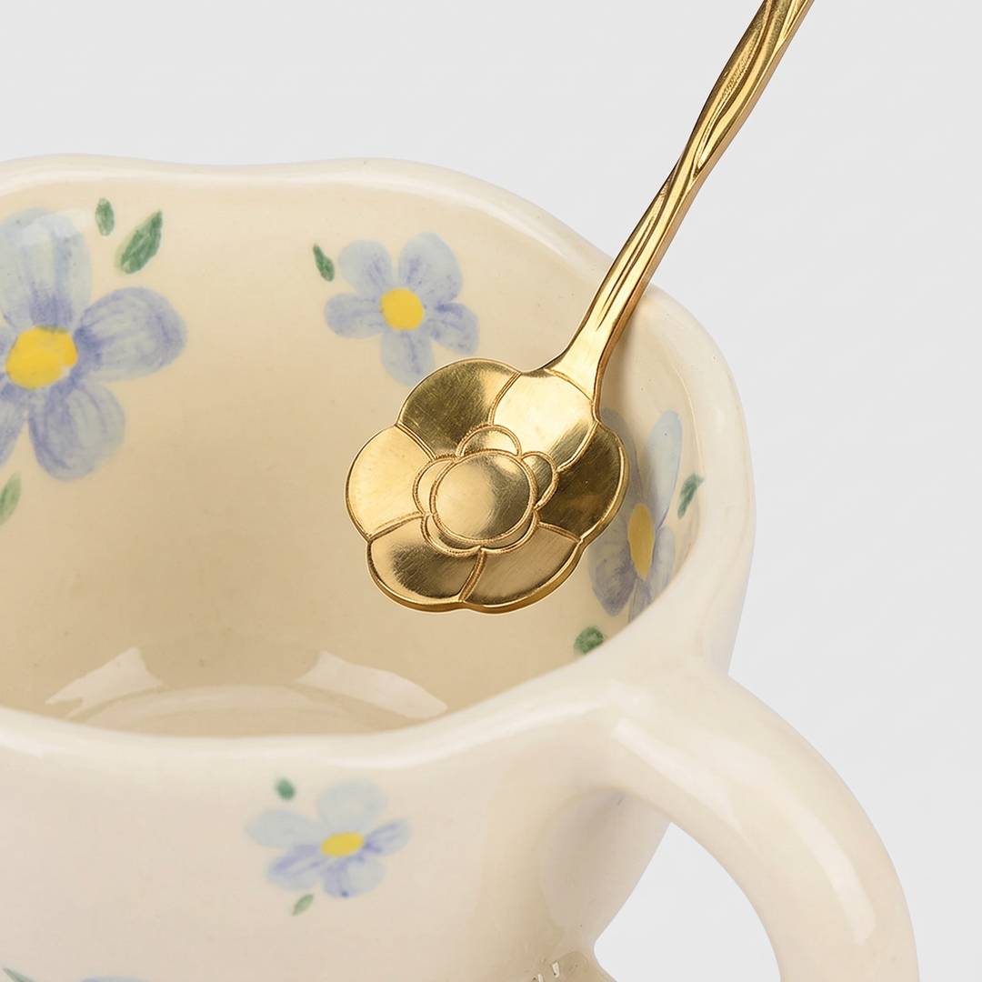 A close-up of a gold-toned flower-shaped spoon resting inside a cream-colored ceramic cup decorated with blue and yellow flowers, on a light gray background.