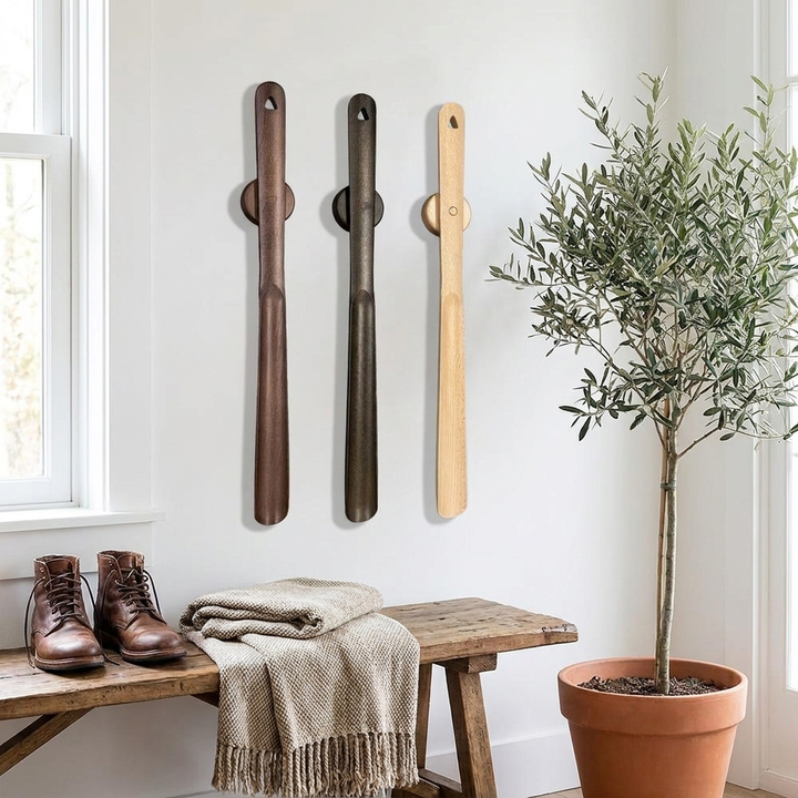 Three wooden shoe tree wall hooks in brown, black, and natural wood tones are displayed on a white wall above a rustic wooden bench with brown leather boots, a folded beige throw blanket, and a potted olive tree nearby.