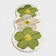 A small decorative mat or plaque with an irregular elongated shape, featuring two green flowers and one cream flower with yellow centers on a light textured background, shown on a plain light gray background.