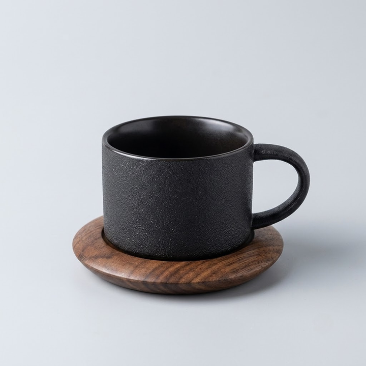 A matte black ceramic coffee mug with a textured finish, resting on a round wooden coaster against a light gray background.