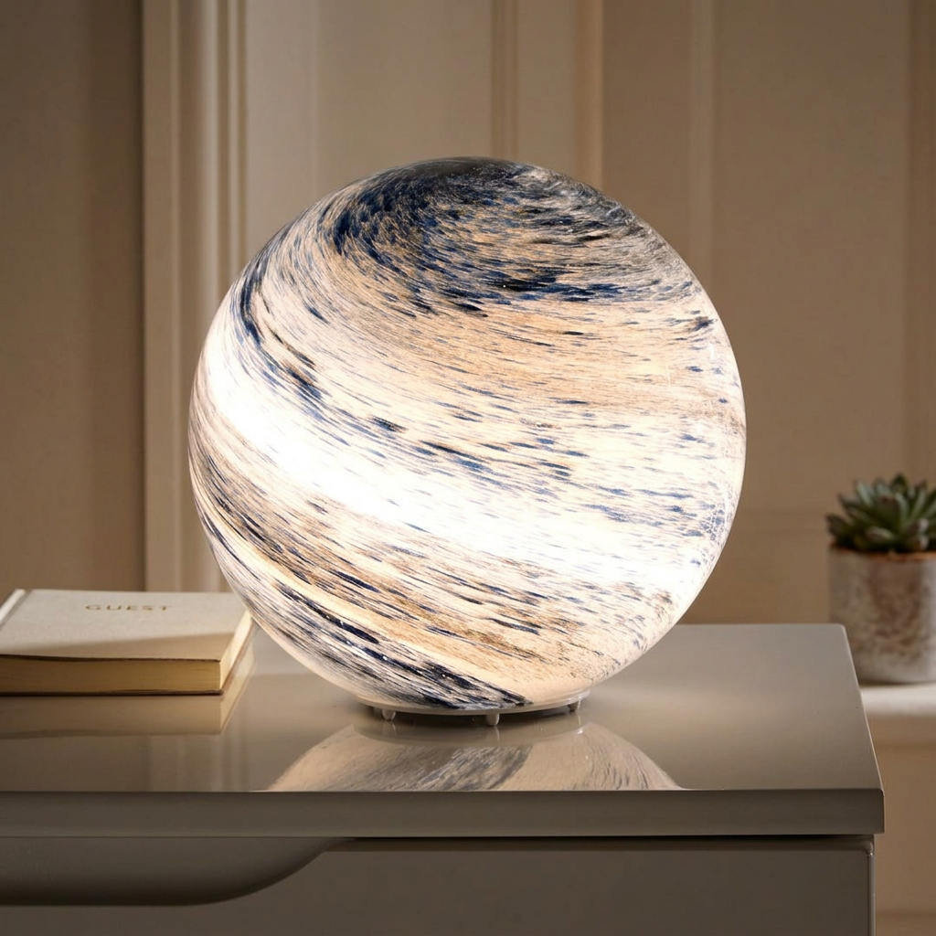 A spherical marbled lamp glowing with warm light, placed on a tabletop in a softly styled interior setting with a small plant and book in the background.