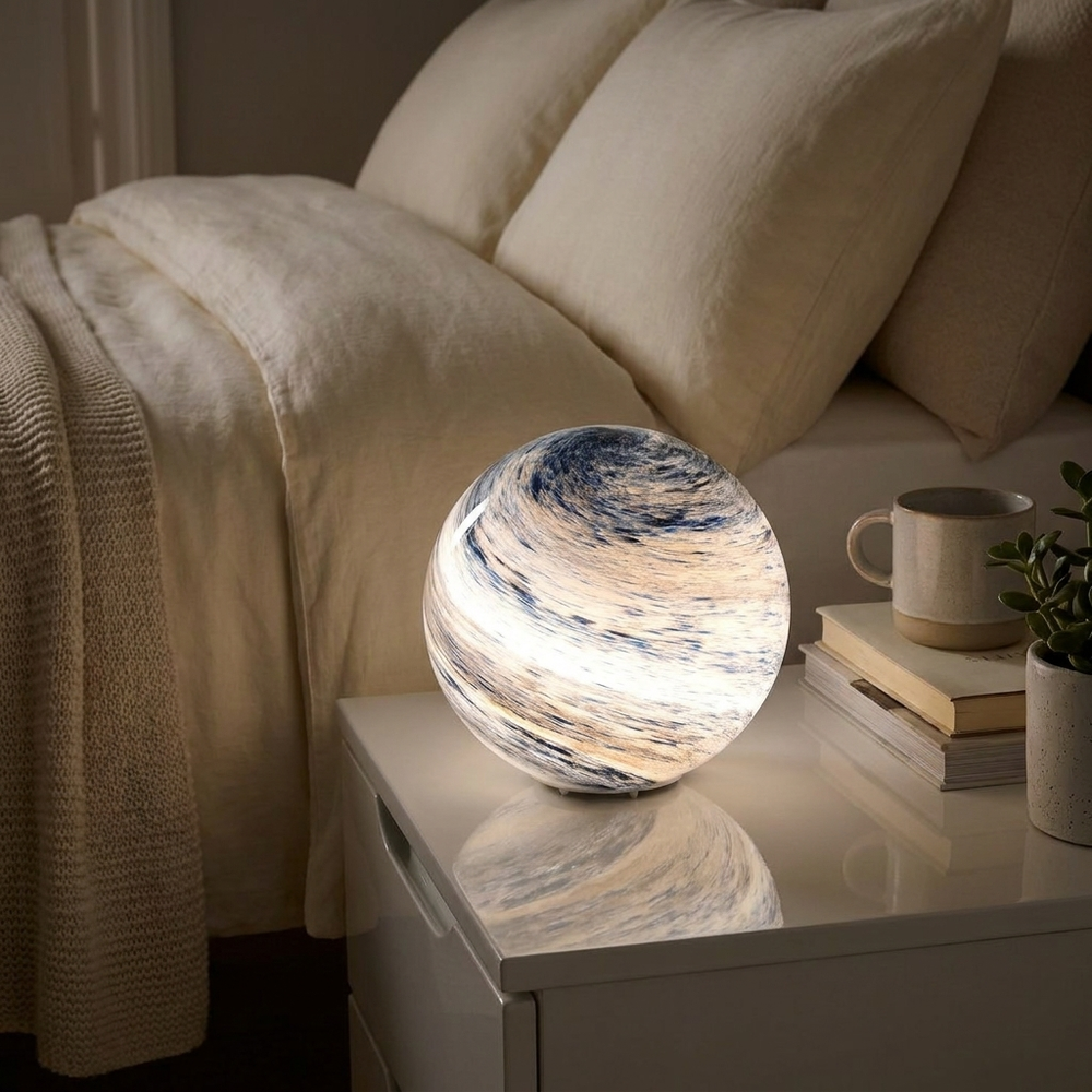 A glowing spherical night light with a marbled blue, cream, and white pattern sits on a white bedside table next to a bed with cream bedding, a mug, and books.
