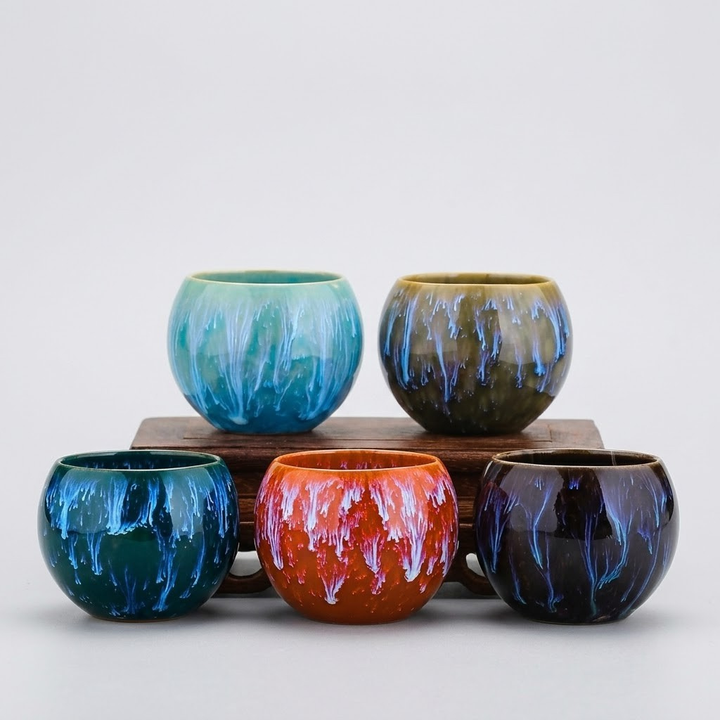 Five small round ceramic cups with marbled, drip-like glaze patterns in assorted colors, arranged on and around a wooden riser against a light gray studio background.